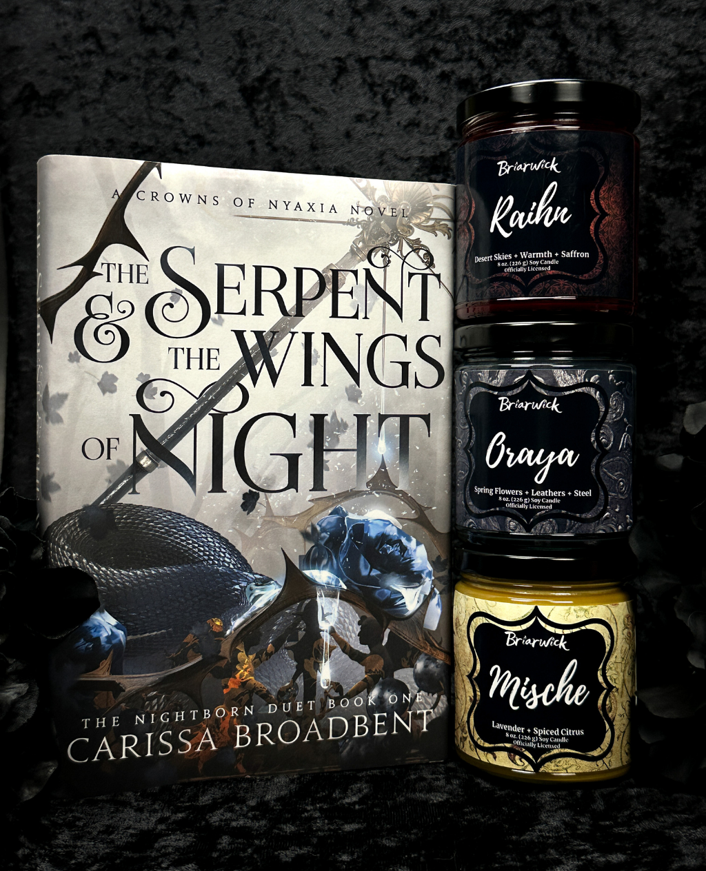 Book titled 'The Serpent & the Wings of Night' by Carissa Broadbent with three branded candles on a dark background.