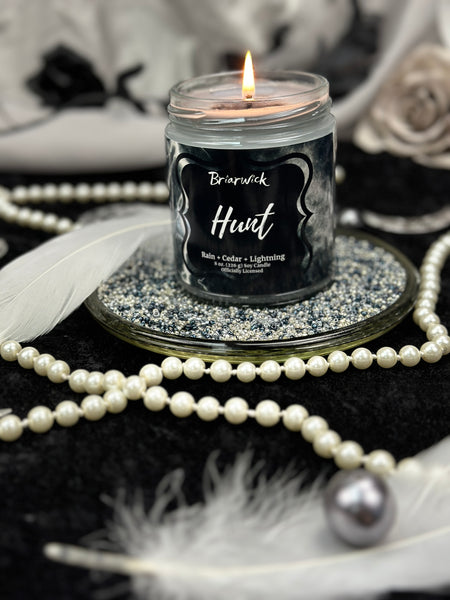Grey candle in a glass jar labeled 'Hunt' with decorative pearls and feathers around it.