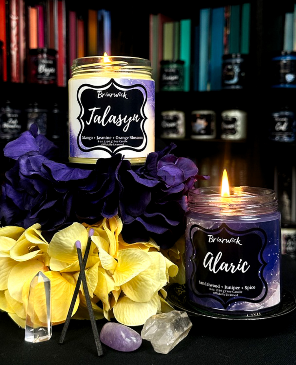 Two lit Talasyn and Alaric candles with labels on a colorful bookshelf background with flowers and crystals.