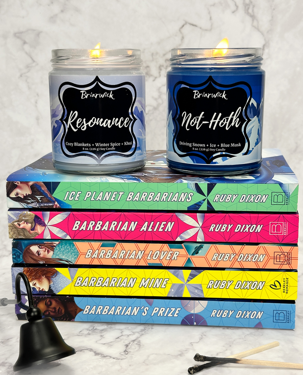 Two lit candles with labels on top of a stack of Ice Planet Barbarians books by Ruby Dixon.