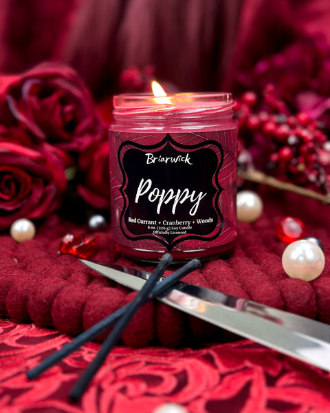 Candle in a jar labeled 'Poppy' surrounded by red roses and berries on a red background