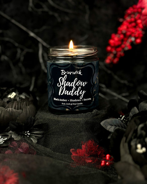 Candle labeled 'Shadow Daddy' in a dark setting with red flowers and black fabric.