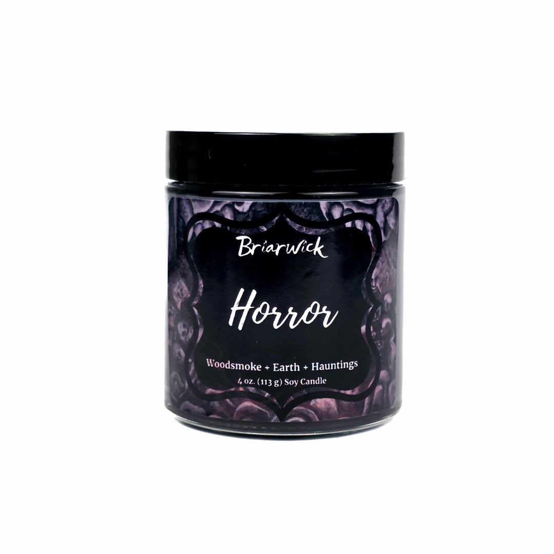 A 4oz soy candle in a clear glass jar with a black lid set on a white background, featuring a dark purple label with gothic horror illustrations and white text that reads 'Briarwick Horror'