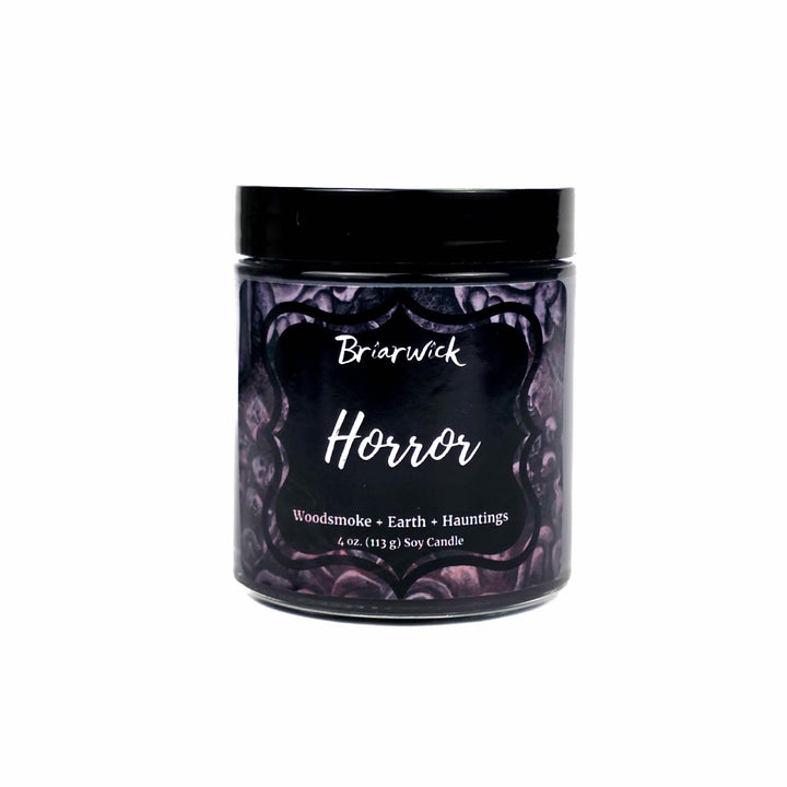 A 4oz soy candle in a clear glass jar with a black lid set on a white background, featuring a dark purple label with gothic horror illustrations and white text that reads 'Briarwick Horror'