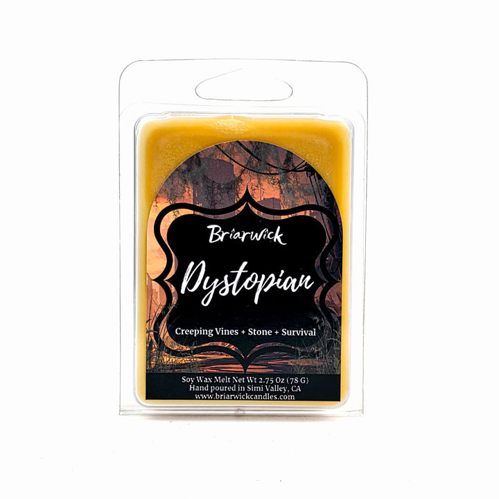 A packaged soy wax melt in a clear plastic clamshell package. The wax itself is a warm golden yellow, and the label features a black background with scent name 'Dystopian'.