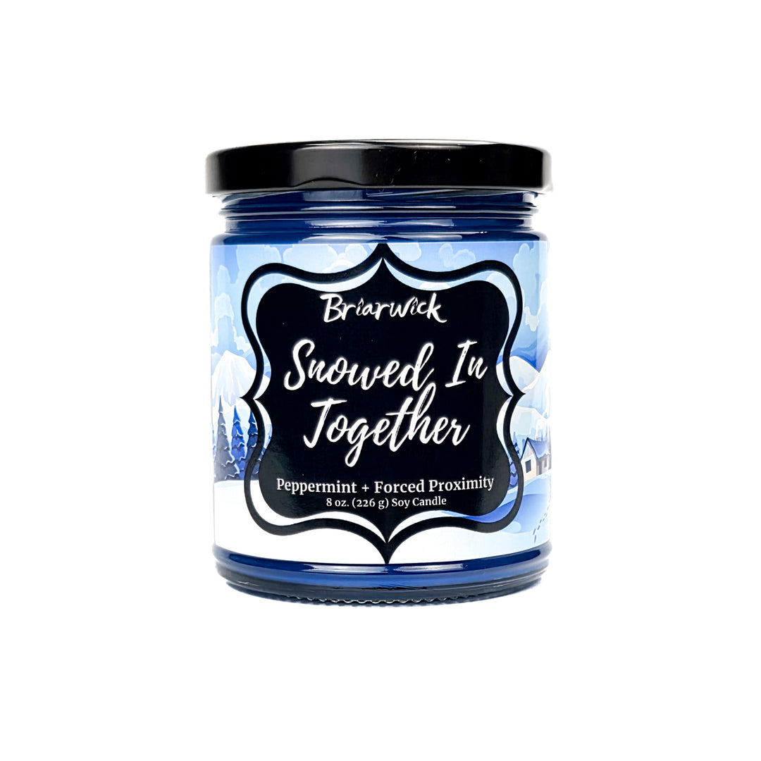 Snowed In Together 8oz colored candle with blue and white snow covered lodge design on a white background